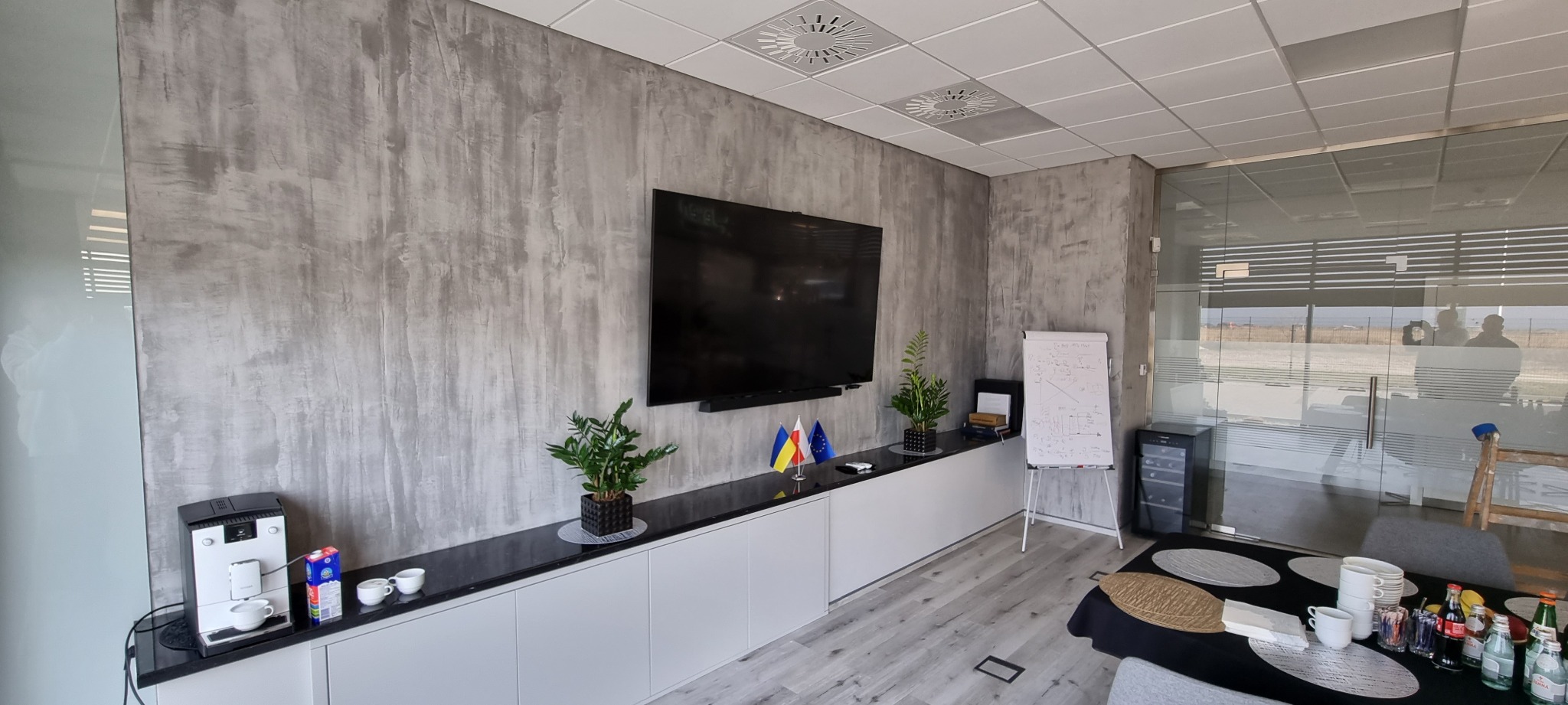 Nowoczesne biuro z ścianą pokrytą szarą fakturą przypominającą beton, telewizor, rośliny doniczkowe, flagi Ukrainy, Polski i Unii Europejskiej, ekspres do kawy i stół konferencyjny z napojami.