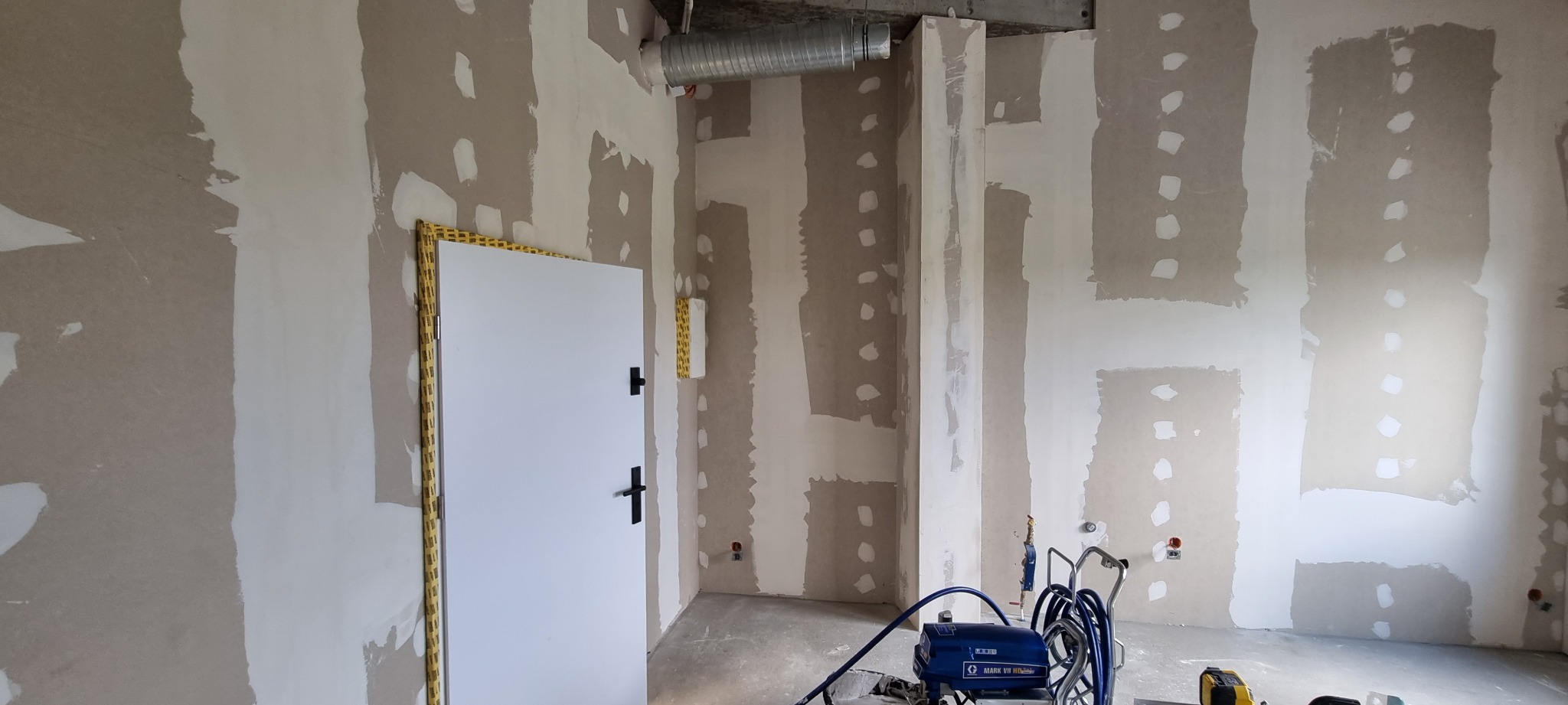 Wnętrze w trakcie prac wykończeniowych, ściany z widocznymi łatami szpachli, białe drzwi z czarnymi klamkami zabezpieczone taśmą, oraz sprzęt do malowania hydrodynamicznego na podłodze.