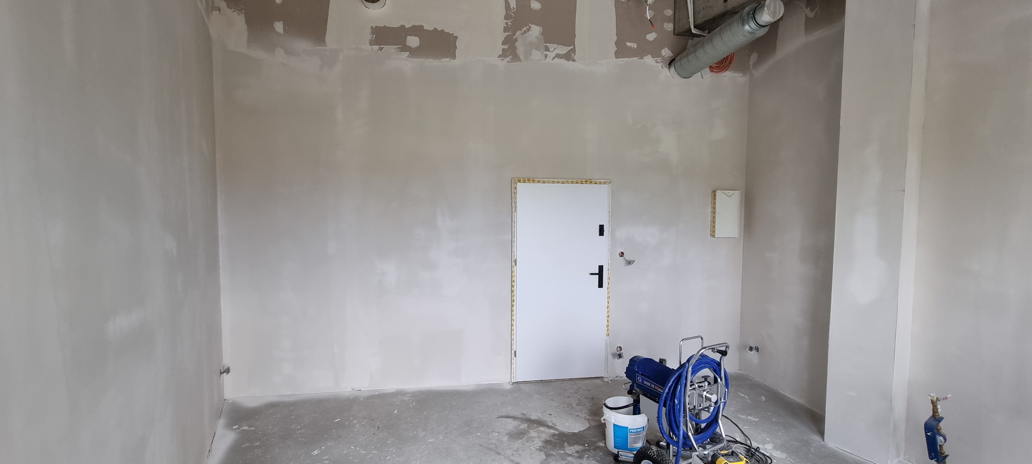 Wnętrze w trakcie prac wykończeniowych: ściany pokryte świeżo nałożoną warstwą, widoczny agregat malarski, białe drzwi z czarną klamką i zabezpieczonymi ościeżnicami, surowy beton na podłodze, rury...