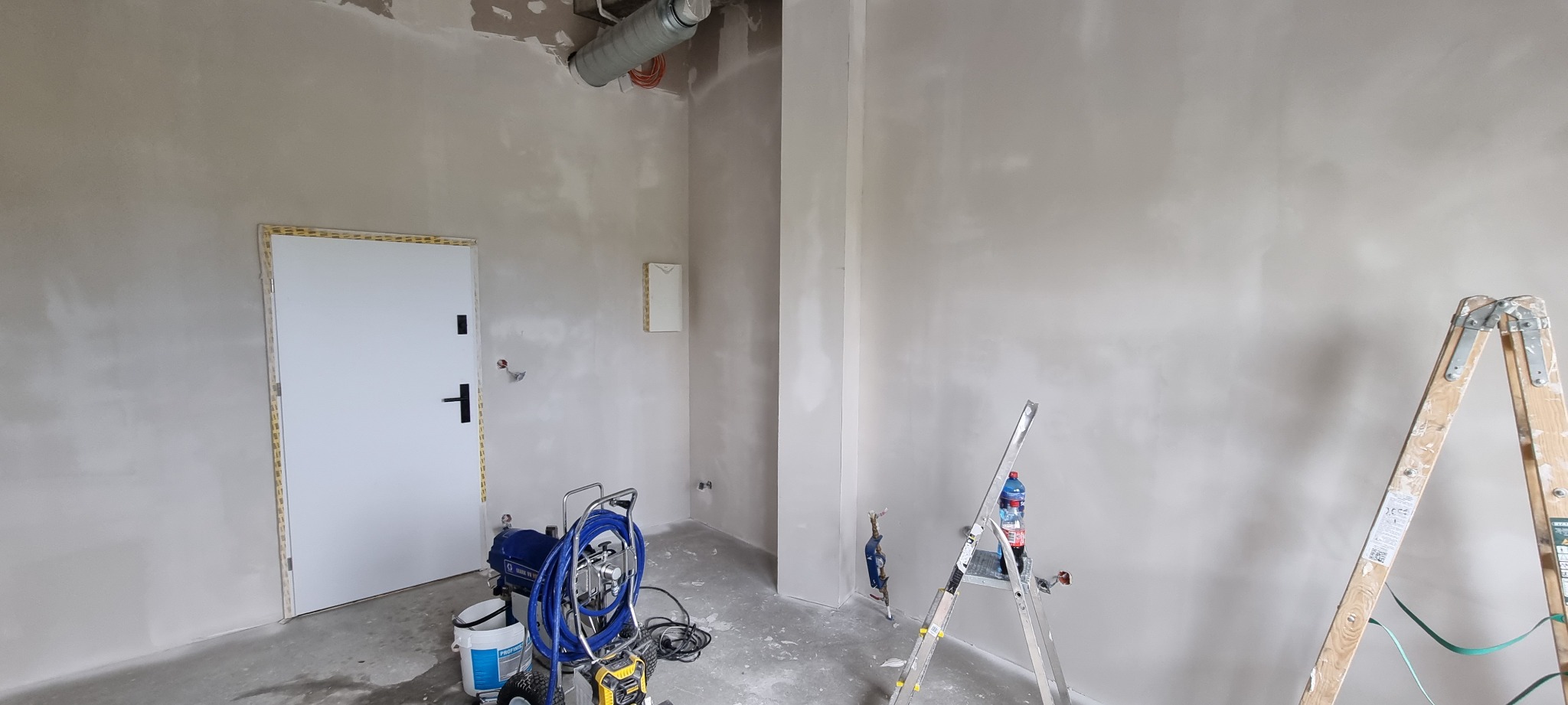 Wnętrze w trakcie szpachlowania: ściany pokryte świeżą warstwą szpachli, widoczna maszyna do szpachlowania natryskowego, drabina z butelką wody, zabezpieczone drzwi.