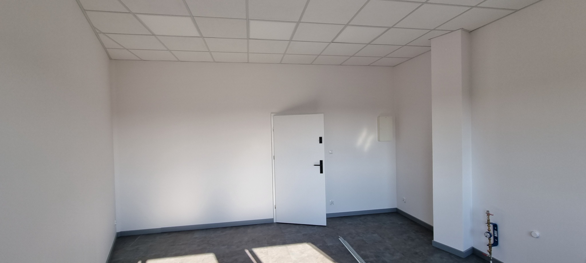 Puste, świeżo wykończone pomieszczenie biurowe z białą ścianą, sufitem kasetonowym i szarą podłogą, widoczne pionowe rury przy ścianie.