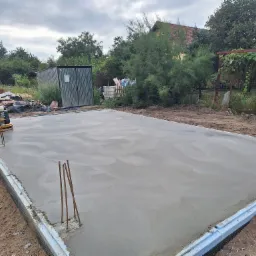 Wygładzona, świeżo wylana płyta fundamentowa z wystającymi prętami zbrojeniowymi, widoczna szlifierka do betonu i niebieska rura kanalizacyjna.