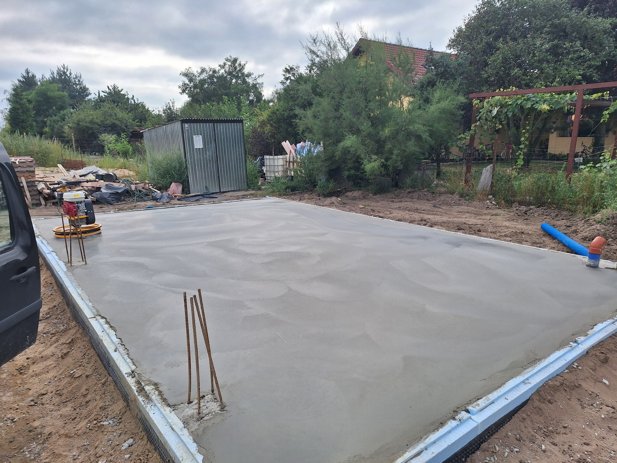Wygładzona, świeżo wylana płyta fundamentowa z wystającymi prętami zbrojeniowymi, widoczna szlifierka do betonu i niebieska rura kanalizacyjna.