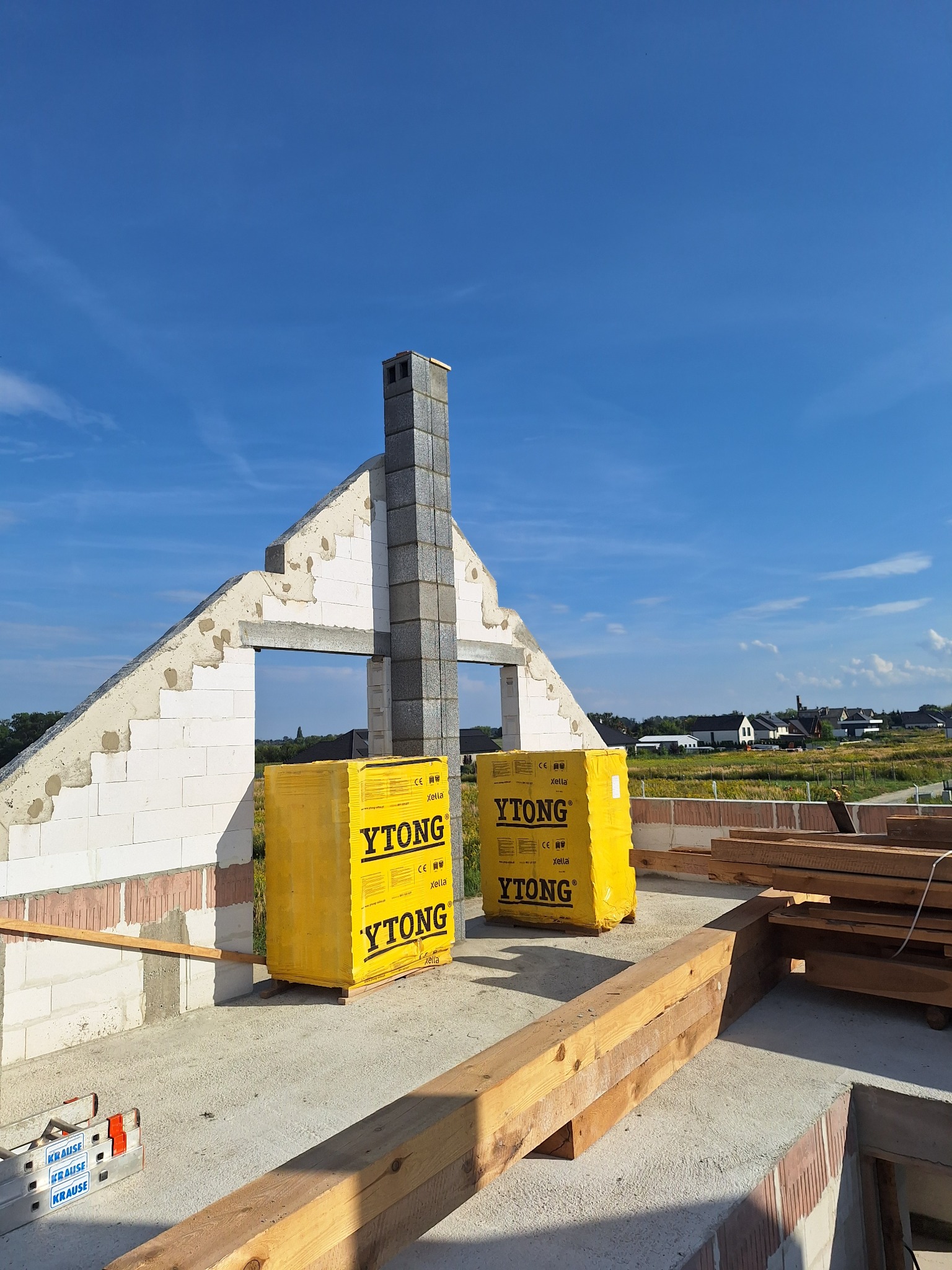 Wznoszenie ścian domu z bloczków Ytong, widoczny komin z szarej cegły, drewniane belki konstrukcyjne i drabina Krause na tle błękitnego nieba i krajobrazu wiejskiego.