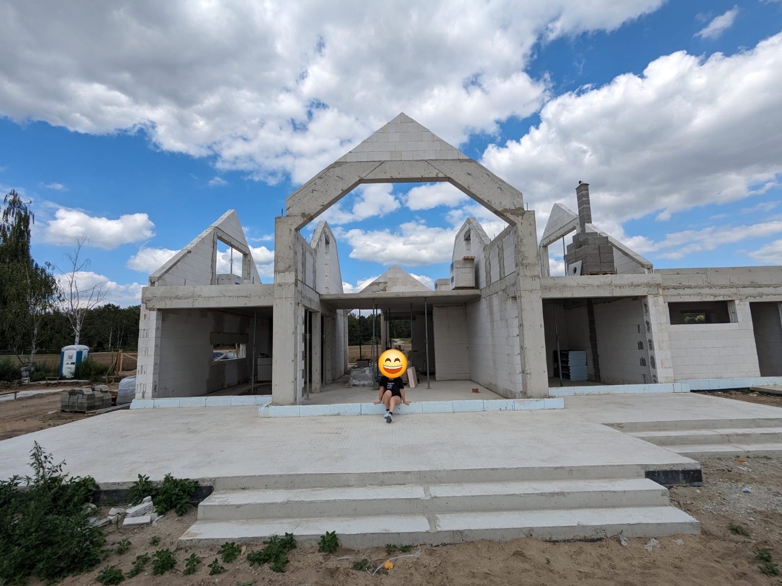 Surowy stan domu z betonu komórkowego z widocznymi ścianami, otworami okiennymi i kominem w trakcie budowy, na pierwszym planie schody i dziecko siedzące na nich, błękitne niebo z chmurami w tle.