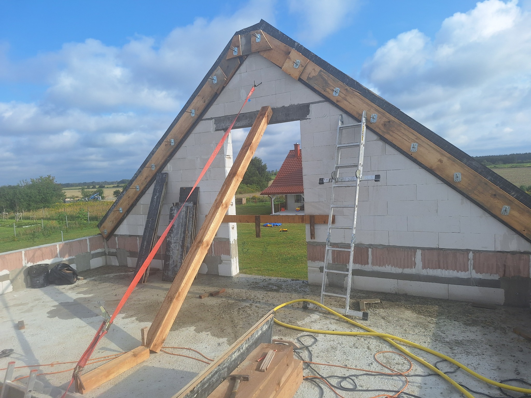 Wznoszenie ścian domu z betonu komórkowego, widoczne drewniane elementy konstrukcyjne dachu, szalunek okienny podtrzymywany czerwoną taśmą, drabina oparta o ścianę, w tle zarys dachu sąsiedniego...