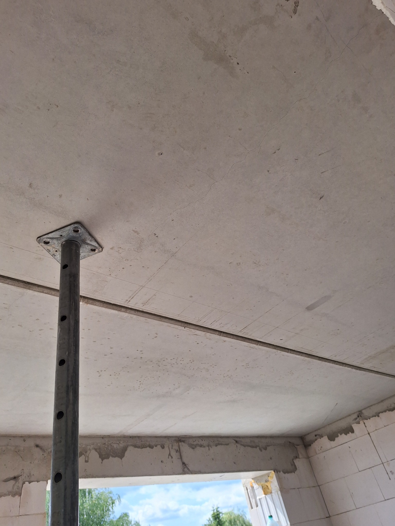 Szary, surowy betonowy strop podparty metalową stemplą budowlaną w trakcie wznoszenia domu z betonu komórkowego, widok na zewnątrz przez otwór okienny.