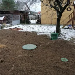 Widok z góry na teren po instalacji przydomowej oczyszczalni ścieków z trzema zielonymi włazami inspekcyjnymi na ubitej ziemi, z pozostałościami śniegu w tle i elementami placu zabaw.