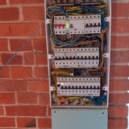 Usługi elektroinstalacyjne i ogólnobudowlane Volt plus - Rozdzielnica elektryczna z bezpiecznikami i przewodami zamontowana na ścianie z czerwonej cegły, widoczne zielone diody LED na jednym z elementów.