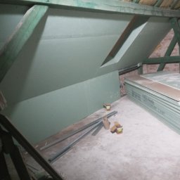 Usługi elektroinstalacyjne i ogólnobudowlane Volt plus - Poddasze w trakcie wykańczania płytami gipsowo-kartonowymi; widoczne drewniane elementy konstrukcyjne, izolacja na suficie, profile metalowe na podłodze oraz stos płyt gipsowych.