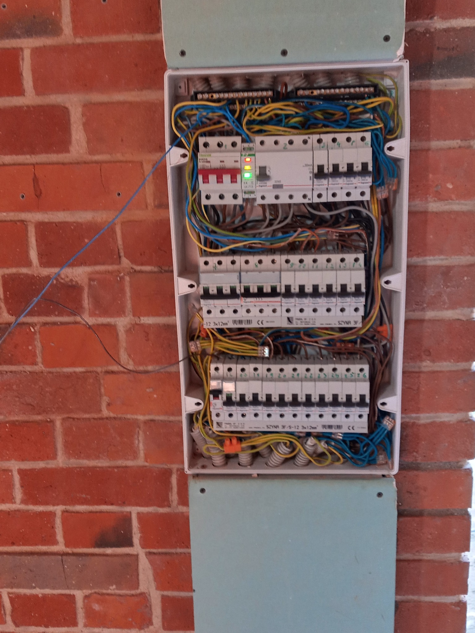 Rozdzielnica elektryczna z bezpiecznikami i przewodami zamontowana na ścianie z czerwonej cegły, widoczne zielone diody LED na jednym z elementów.
