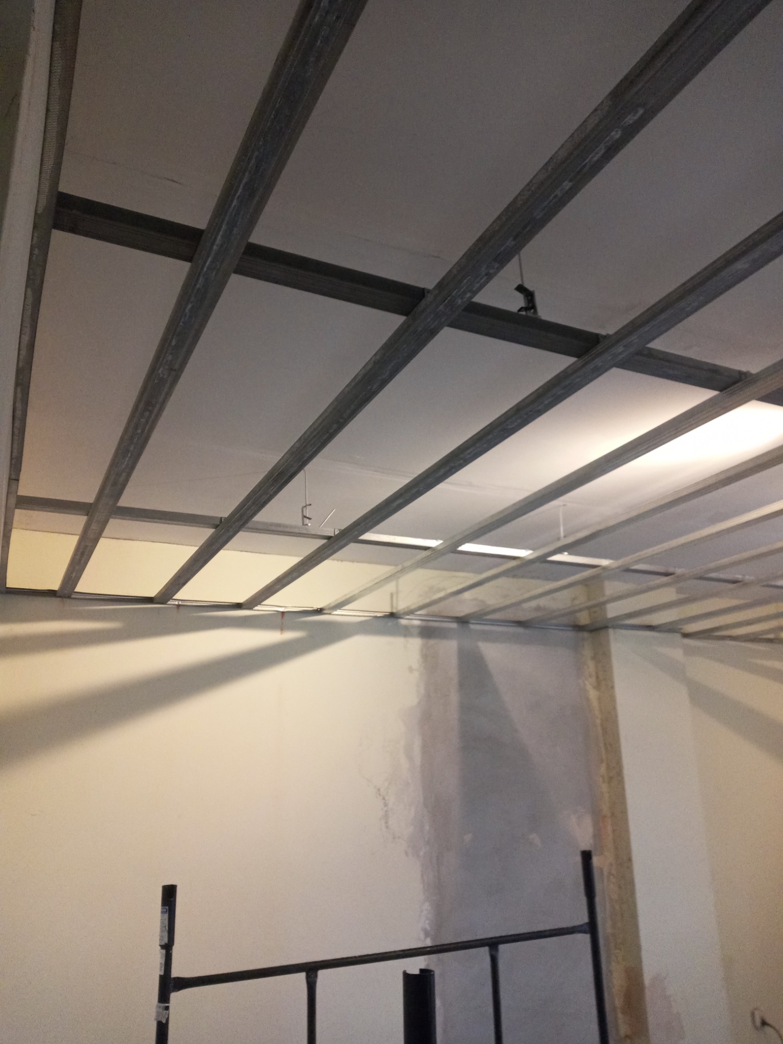 Stelaż sufitowy z profili metalowych, częściowo wypełniony płytami, widok z dołu na sufit w trakcie montażu, surowe ściany w tle.