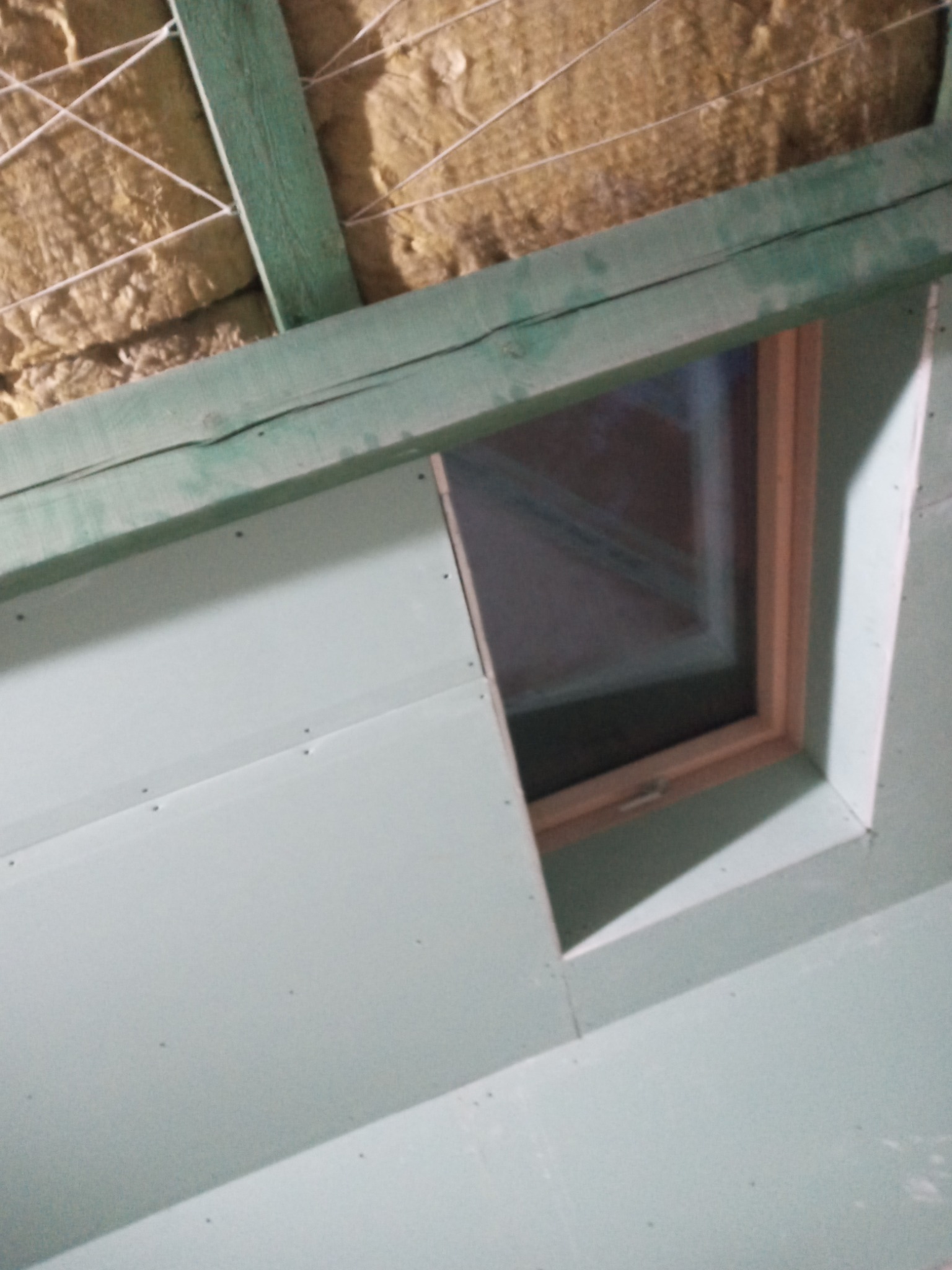 Montaż płyty gipsowo-kartonowej wokół okna dachowego, widoczna izolacja wełną mineralną i drewniana konstrukcja nośna.