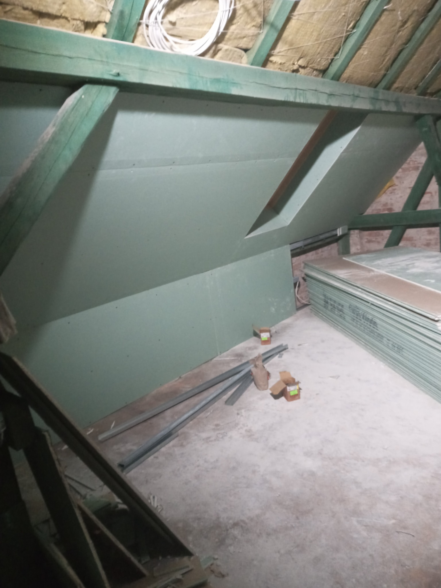 Poddasze w trakcie wykańczania płytami gipsowo-kartonowymi; widoczne drewniane elementy konstrukcyjne, izolacja na suficie, profile metalowe na podłodze oraz stos płyt gipsowych.