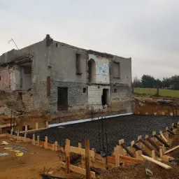 Przygotowanie zbrojenia fundamentów na placu budowy, obok pozostałości starego, częściowo zburzonego budynku z cegły i betonu.