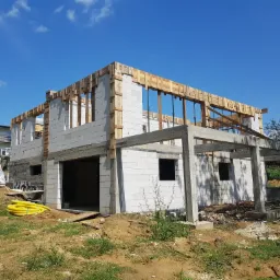 Wznoszony dwukondygnacyjny dom z betonu komórkowego, z drewnianym szalunkiem na stropie i betonowym zadaszeniem tarasu, widoczne żółte rury i betoniarka.