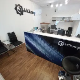 Wnętrze punktu usługowego Mckomp z ladą recepcyjną z logo, krzesłami dla klientów i widocznym stanowiskiem pracy za szybą.