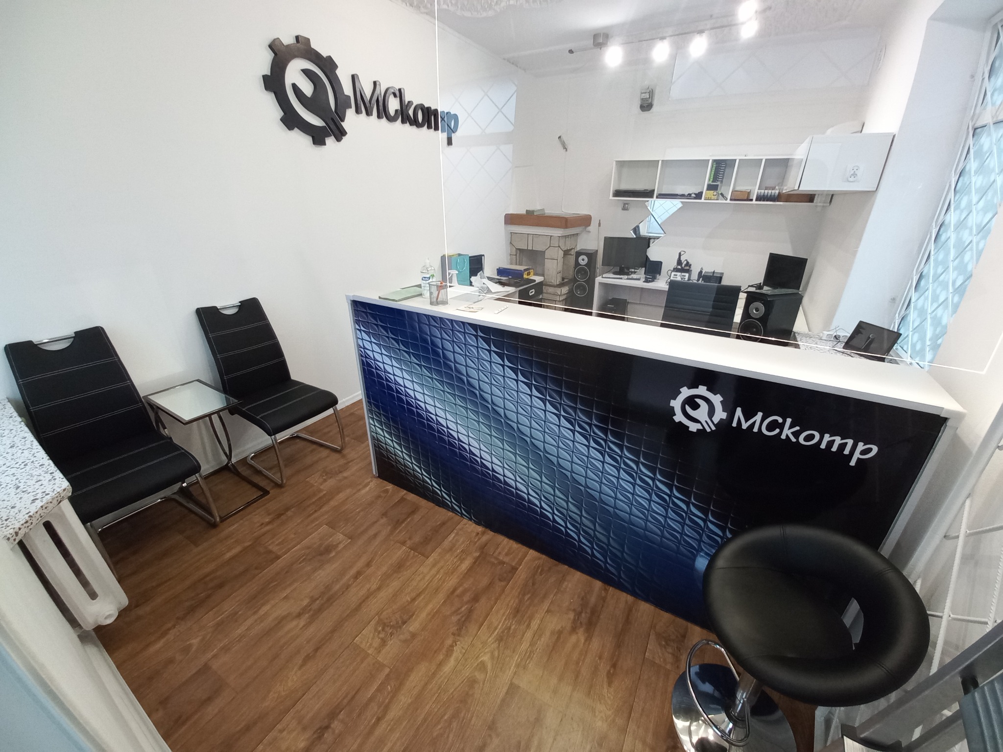 Wnętrze punktu usługowego Mckomp z ladą recepcyjną z logo, krzesłami dla klientów i widocznym stanowiskiem pracy za szybą.
