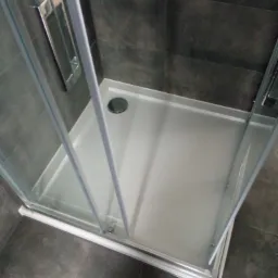 Nowoczesny brodzik z kabiną prysznicową o szklanych drzwiach i chromowanych uchwytach, wyłożony ciemnymi płytkami.