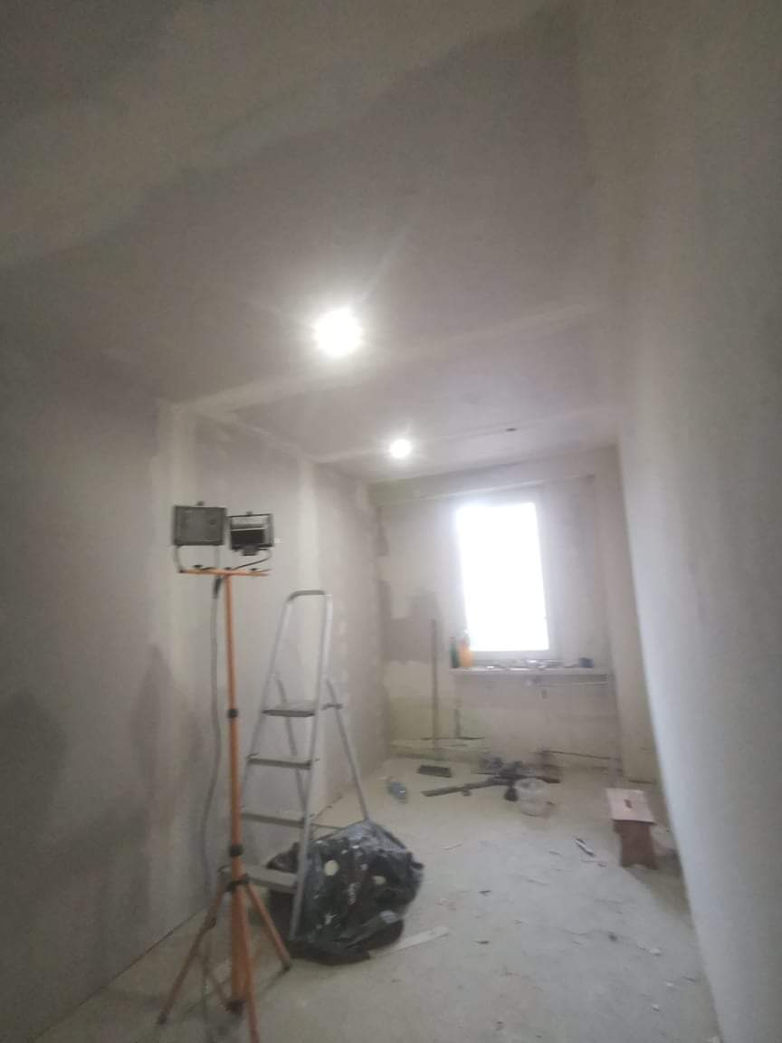 Pokój w trakcie remontu: ściany po szpachlowaniu, drabina, lampa halogenowa na statywie, okno z parapetem, widoczne ślady prac remontowych na podłodze.