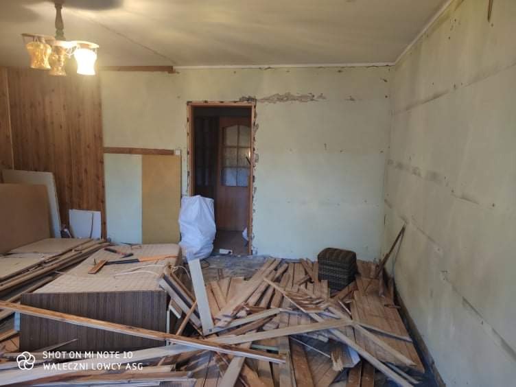 Pokój w trakcie demontażu, widoczne odsłonięte ściany, resztki podłogi, elementy drewniane i meblowe, przygotowania do dalszych prac remontowych.