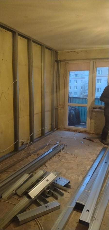 Wnętrze pokoju w trakcie remontu, widoczne metalowe profile do płyt kartonowo-gipsowych przymocowane do ściany, na podłodze leżą dodatkowe profile, okno z widokiem na budynek.