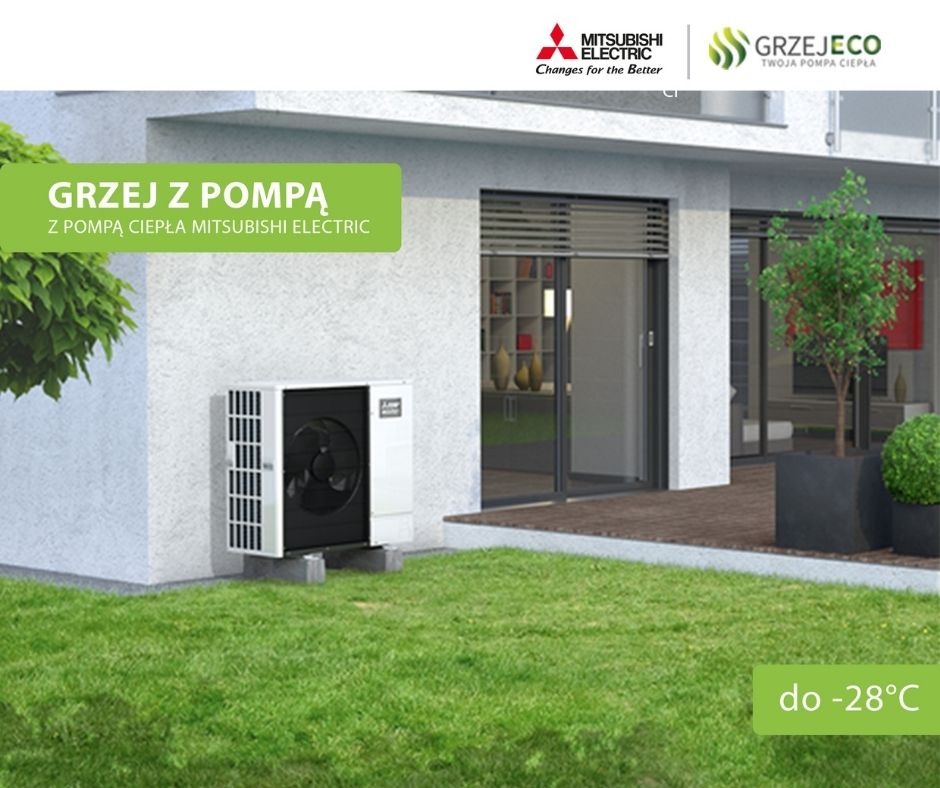 Zewnętrzna jednostka pompy ciepła Mitsubishi Electric, zamontowana przy ścianie budynku z zielonym trawnikiem i dekoracyjnymi krzewami, widoczna z boku, z informacją o działaniu do -28 stopni...