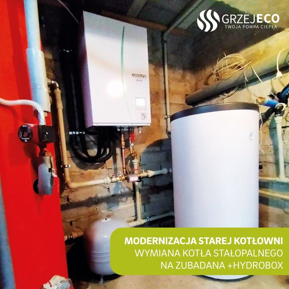 Zmodernizowana kotłownia: biały zasobnik ciepłej wody i wiszący moduł grzewczy Ecodan w surowym wnętrzu z ceglaną ścianą. Widoczna czerwona obudowa pompy obiegowej oraz instalacja rurowa.