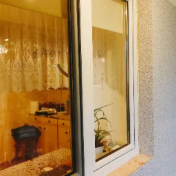 Fragment elewacji z oknem PCV, widoczna struktura tynku baranka w odcieniach szarości i beżu, żółta taśma maskująca przy oknie, parapet z plamami, w oknie odbicie wnętrza z zasłonką i rośliną...