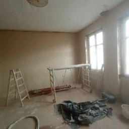Pokój w trakcie remontu, ściany pokryte świeżym tynkiem, widoczne rusztowanie, drabina, narzędzia i materiały budowlane na podłodze, otwór w suficie z wystającymi kablami.