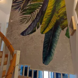 Ściana w klatce schodowej z tapetą w tropikalne liście, widok od dołu schodów z drewnianymi stopniami i białymi barierkami, oświetlona lampkami choinkowymi.