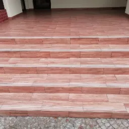 Schody zewnętrzne wyłożone płytkami imitującymi drewno, z subtelnym wzorem słojów i jasnymi fugami, prowadzące do wejścia budynku z ceglanym obramowaniem.