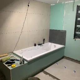 Wanna obudowana płytami kartonowo-gipsowymi, częściowo wyłożona płytkami, widoczne narzędzia i wiadro z zaprawą, podłoga w trakcie układania płytek.