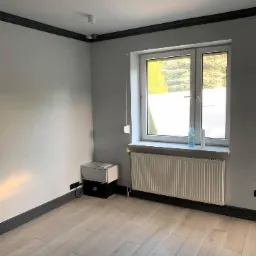 Pusty pokój z jasnymi panelami podłogowymi, szarymi ścianami i ciemnymi listwami przypodłogowymi oraz sufitowymi. Białe okno z grzejnikiem pod spodem. Widoczna szafka przy ścianie z wystającymi...