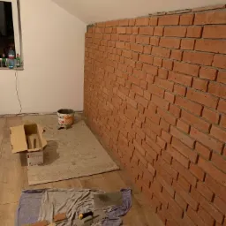 Montaż cegieł dekoracyjnych na ścianie w pomieszczeniu ze skosem, widoczny kubeł z zaprawą, narzędzia i kartony na podłodze.