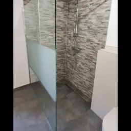 Nowoczesna kabina prysznicowa typu walk-in z częściowo matową szybą, deszczownicą i odpływem liniowym w podłodze, wykończona płytkami imitującymi kamień w odcieniach szarości i beżu, z widoczną...