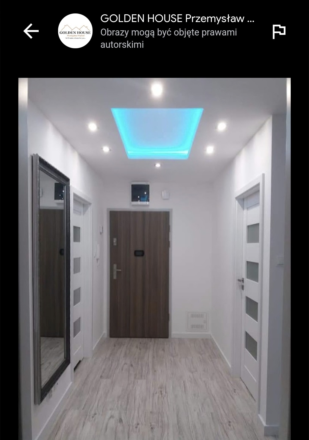 Korytarz w mieszkaniu z białymi ścianami, drewnianą podłogą, lustrem w czarnej ramie i podwieszanym sufitem z oświetleniem LED w kolorze turkusowym.