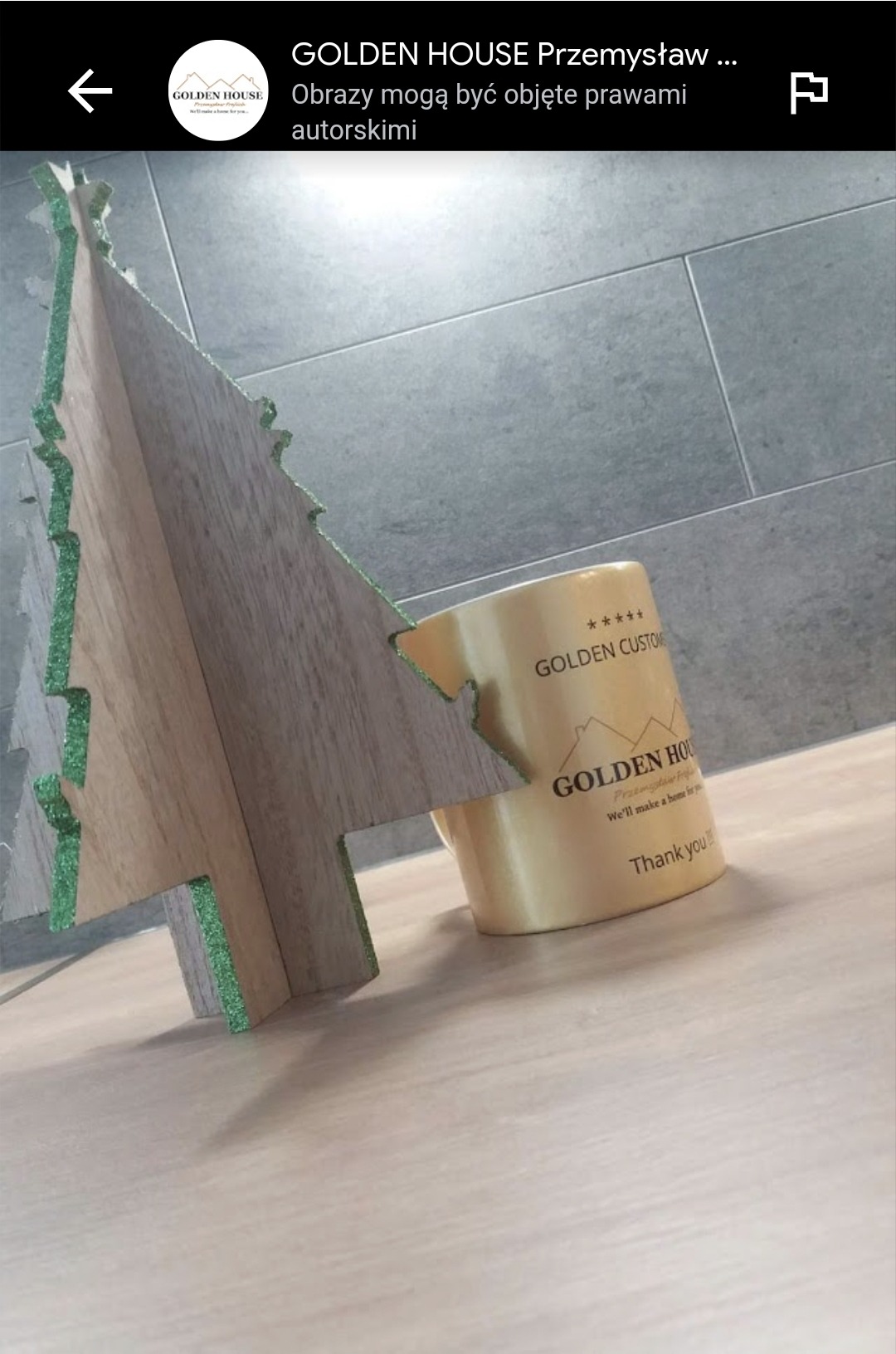 Dekoracyjna choinka z płyty drewnopodobnej z zielonymi brokatowymi krawędziami, ustawiona na podłodze z paneli, obok kubek z logo Golden House.