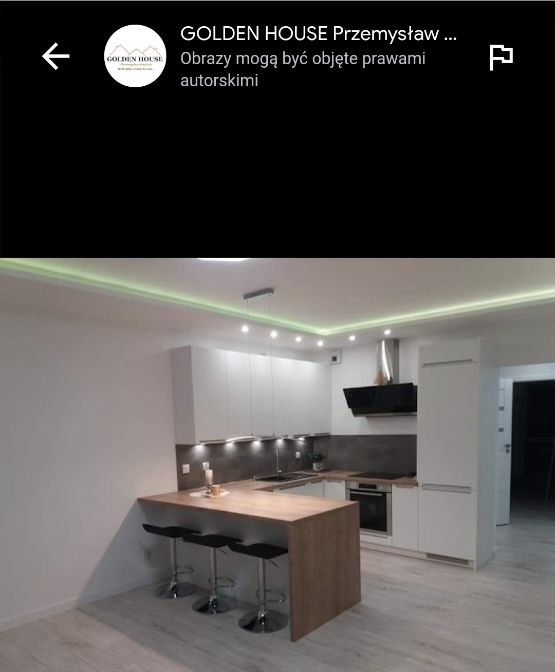 Nowoczesna, jasna kuchnia z białymi szafkami i drewnianym blatem, oświetlona listwą LED pod sufitem, z trzema czarnymi hokerami przy blacie.