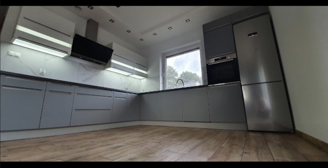 Nowoczesna kuchnia z szarymi szafkami, drewnianą podłogą i oświetleniem LED pod szafkami wiszącymi, widok na okno z widokiem na zewnątrz.