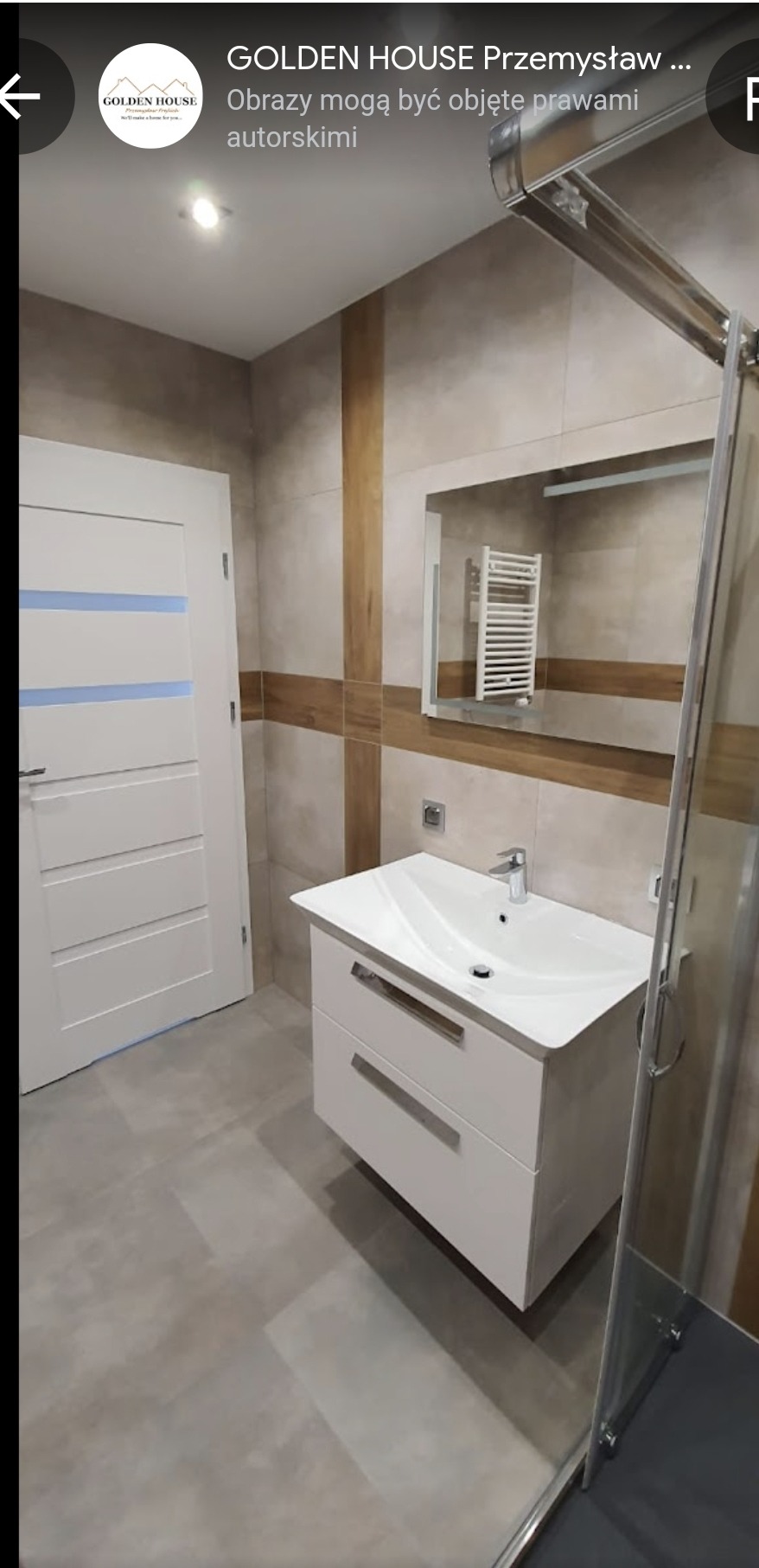 Wykończona łazienka z białą szafką pod umywalką, lustrem, grzejnikiem drabinkowym odbitym w lustrze, szklaną kabiną prysznicową i drzwiami z poziomymi paskami w kolorze niebieskim.