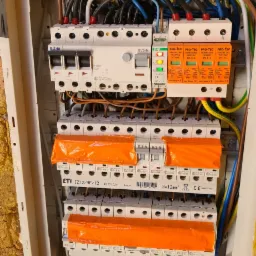 Rozdzielnica elektryczna z zabezpieczeniami przepięciowymi PRO-TEC, bezpiecznikami EATON i ETI, widoczne przewody w różnych kolorach oraz pomarańczowe osłony na dolnych bezpiecznikach.