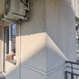 Zewnętrzna jednostka klimatyzacji zamontowana na balkonie, z widocznymi przewodami i odpływem skroplin do czarnego pojemnika, obok okna z kartonowym pudełkiem w tle.