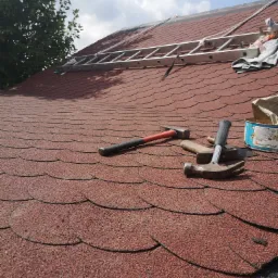 Czerwony dach pokryty gontem bitumicznym, z narzędziami dekarskimi: młotki, puszka po kleju i drabina oparta o dach.