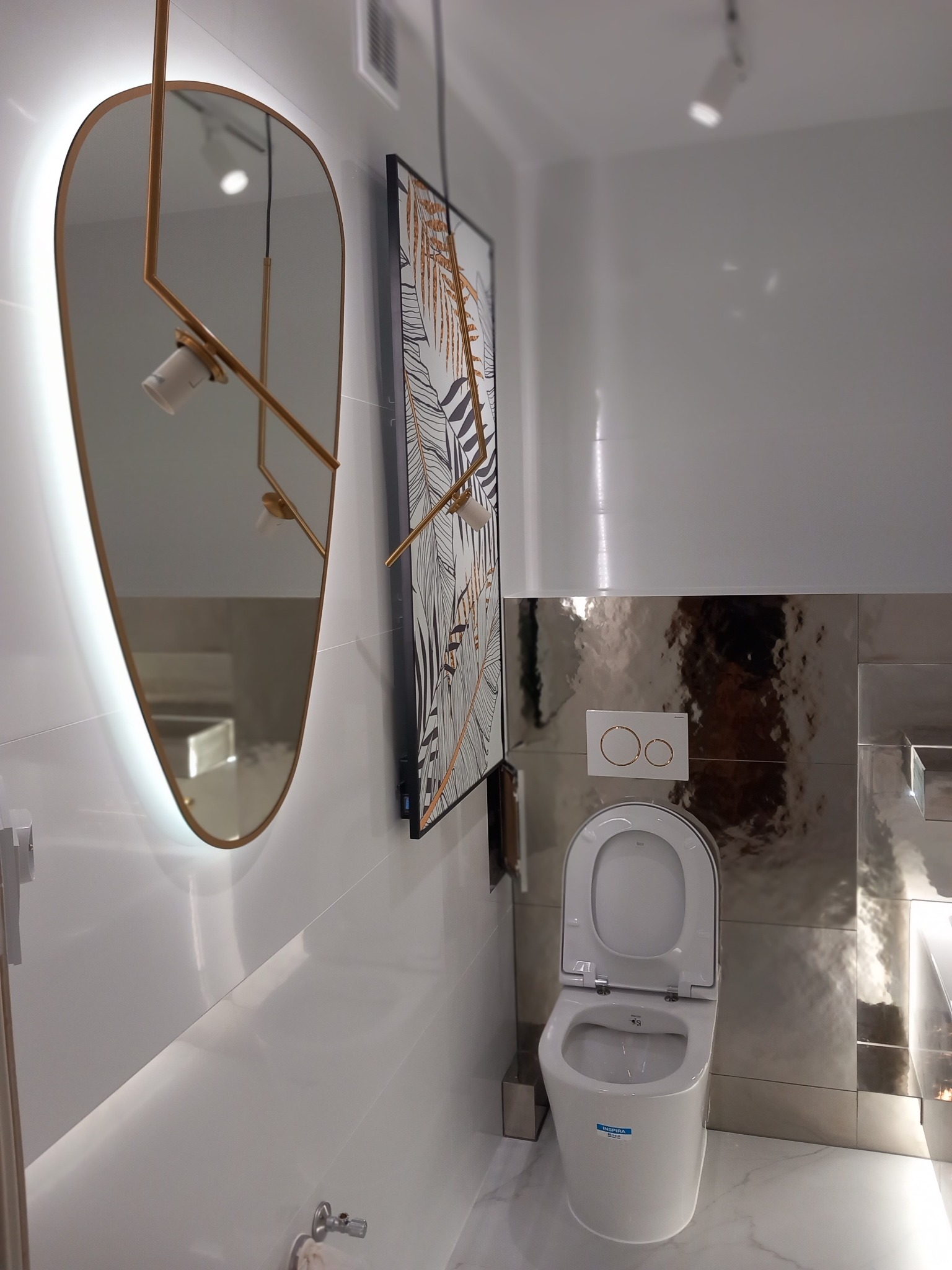 Nowoczesna łazienka z wiszącą toaletą Roca Inspira, lustrem z podświetleniem LED i dekoracyjną grafiką w czarno-złote liście, na tle błyszczących, srebrnych płytek.