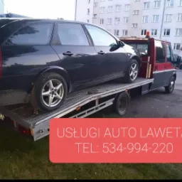 Czarny samochód kombi na platformie lawety, czerwono-szara kabina lawety na tle bloku mieszkalnego.