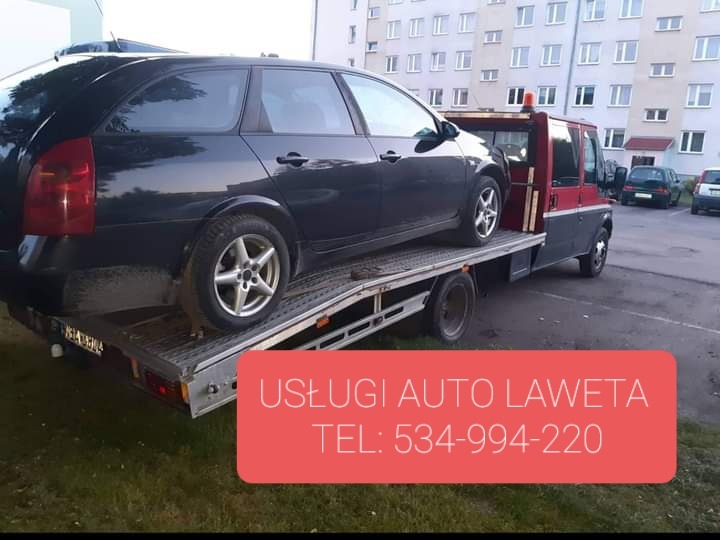 Czarny samochód kombi na platformie lawety, czerwono-szara kabina lawety na tle bloku mieszkalnego.