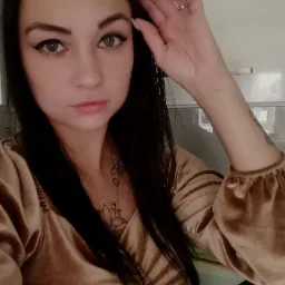 Portret kobiety z ciemnymi włosami, w złotej bluzce z bufiastymi rękawami, z tatuażami na dekolcie i ramieniu, patrzącej w obiektyw, z ręką uniesioną do czoła, na tle jasnej ściany.