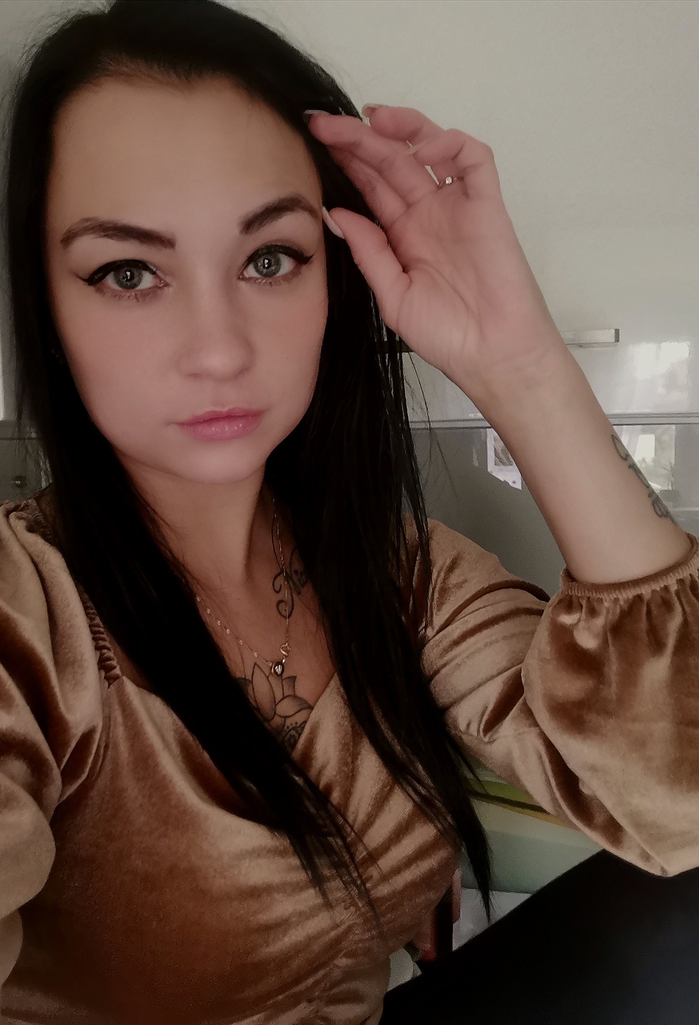 Portret kobiety z ciemnymi włosami, w złotej bluzce z bufiastymi rękawami, z tatuażami na dekolcie i ramieniu, patrzącej w obiektyw, z ręką uniesioną do czoła, na tle jasnej ściany.