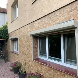Beżowe rolety zewnętrzne zamontowane na oknach domu z elewacją imitującą kamień, widok z boku na budynek, z widocznymi donicami z kwiatami i fragmentem zadaszenia z roślinnością.